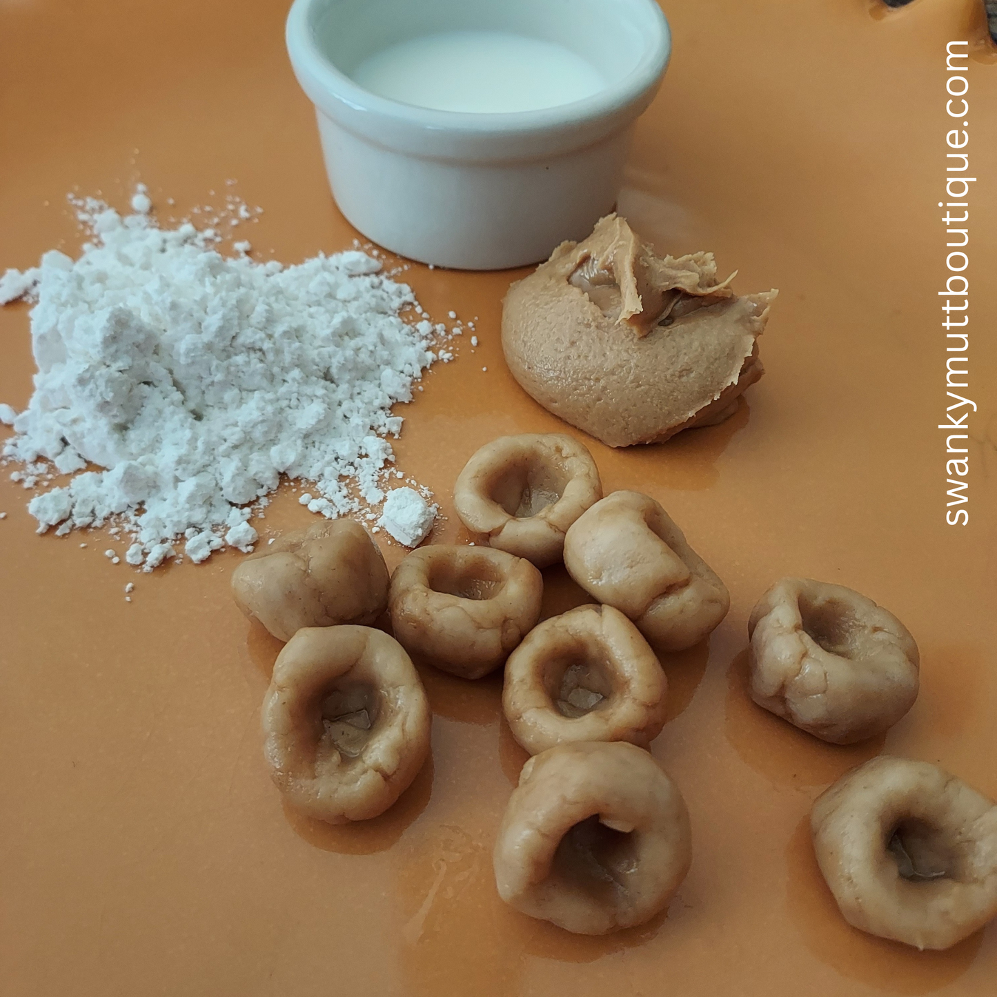 Dog treats with a scoop of flour and a small container of milk on an orange surface, branded 'swankymuttboutique.com'.
