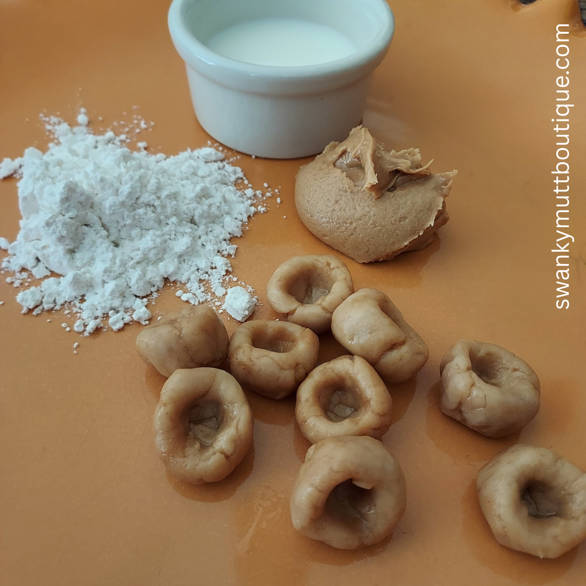 Dog treats with a scoop of flour and a small container of milk on an orange surface, branded 'swankymuttboutique.com'.