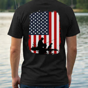 Black t-shirt with American flag and silhouette of a person fishing on the back, worn by a person standing by a body of water.