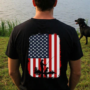 Person wearing a black t-shirt with an American flag design and a fisherman by a lake