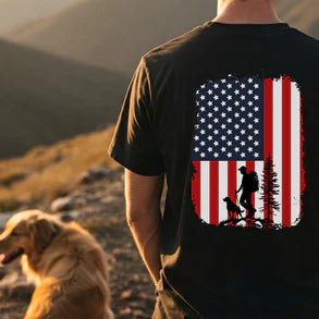 Person wearing a black t-shirt with an American flag design and a dog walking silhouette, standing in a mountainous landscape.