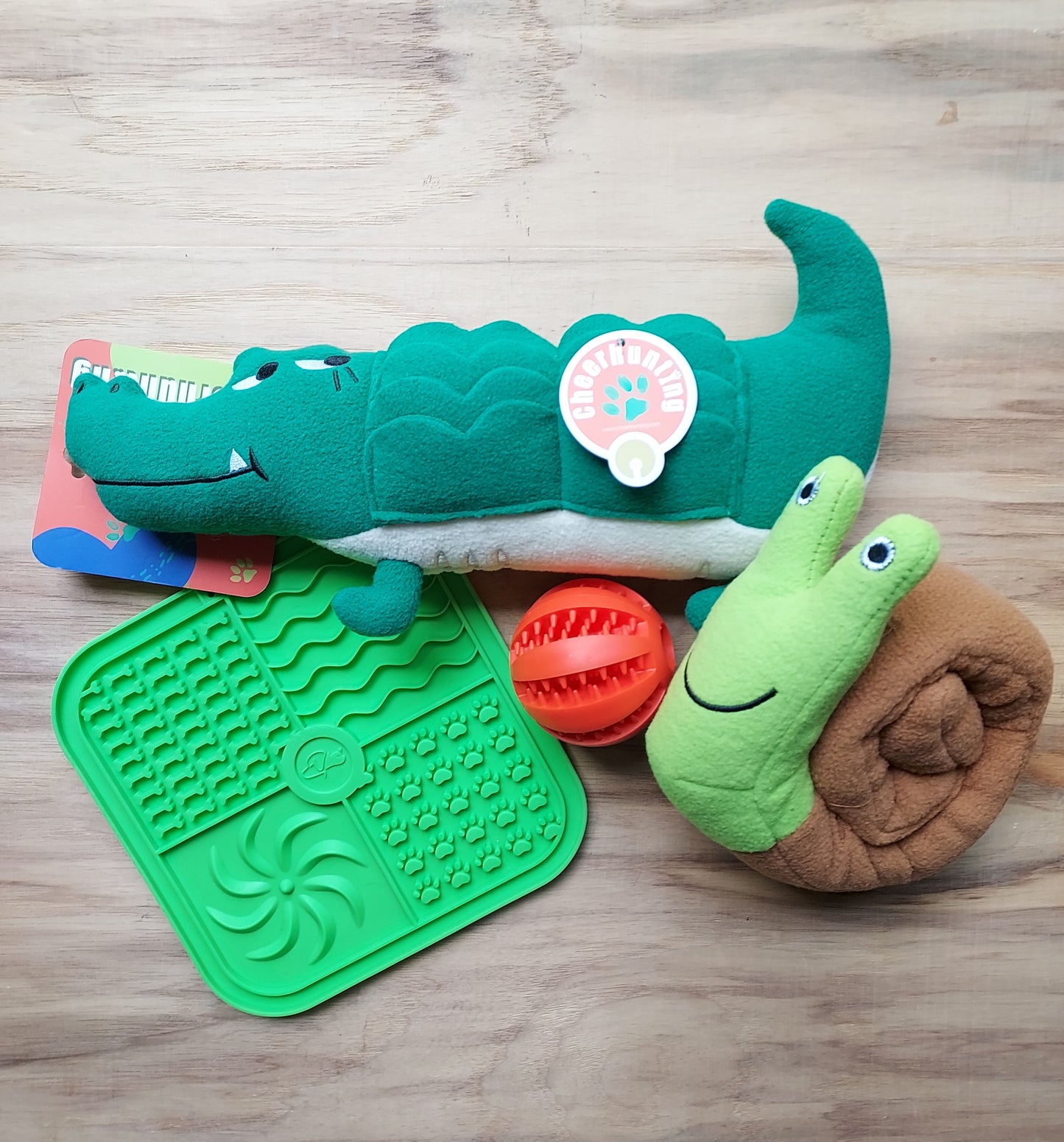 Set of dog toys including a green alligator, red ball, and green snail on a wooden surface.